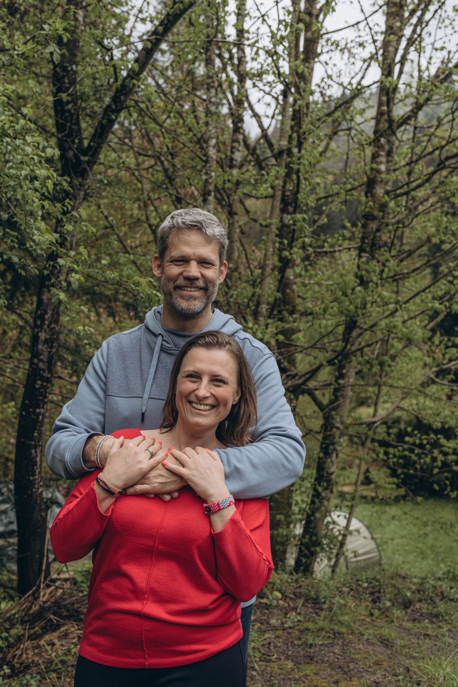 Markus steht hinter Olga und legt die Arme um sie, beide lächeln, im Hintergrund Wald